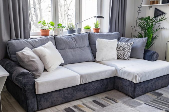 Canapé convertible d'angle bicolore gris et blanc avec coussins décoratifs près d'une fenêtre fleurie