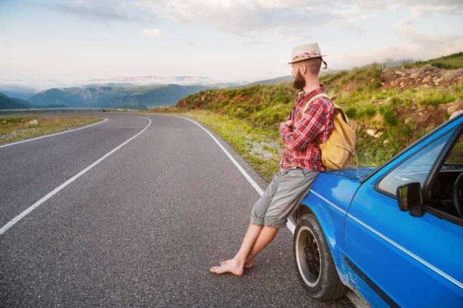 Un homme barbu avec un sac à dos s'appuie sur une voiture bleue face à une route de montagne, prêt à partir en voyage.