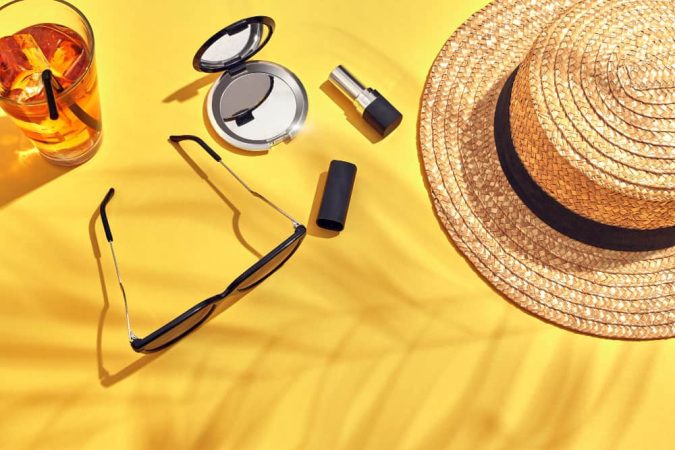 Nature morte sur fond jaune vif présentant un chapeau de paille, du maquillage et des accessoires été créateur soleil