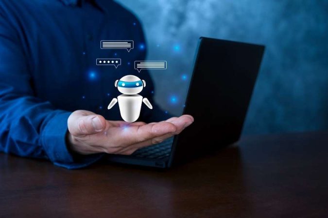 Un homme tient un petit robot blanc virtuel au-dessus de sa main ouverte, à côté d'un ordinateur portable noir.