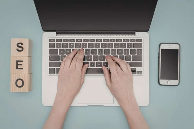 Vue du dessus de mains tapant sur le clavier d'un ordinateur portable, entouré de blocs en bois affichant SEO et d'un smartphone.