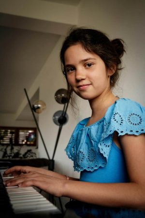 Une jeune fille souriante en haut bleu joue du piano dans une pièce lumineuse.