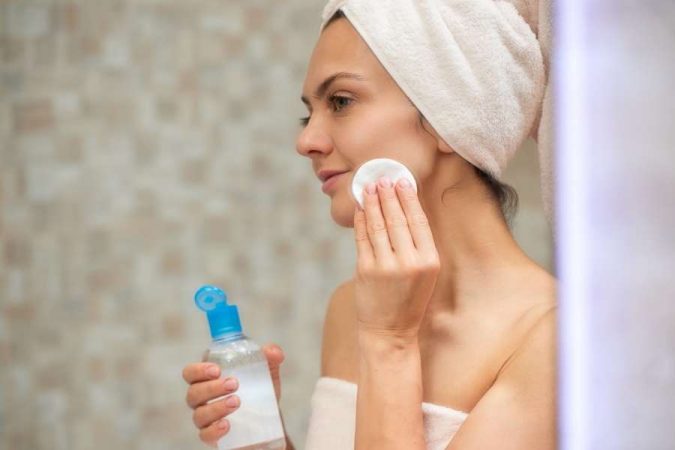 Une femme avec une serviette sur la tête applique un soin de sa routine de soins avec un coton.