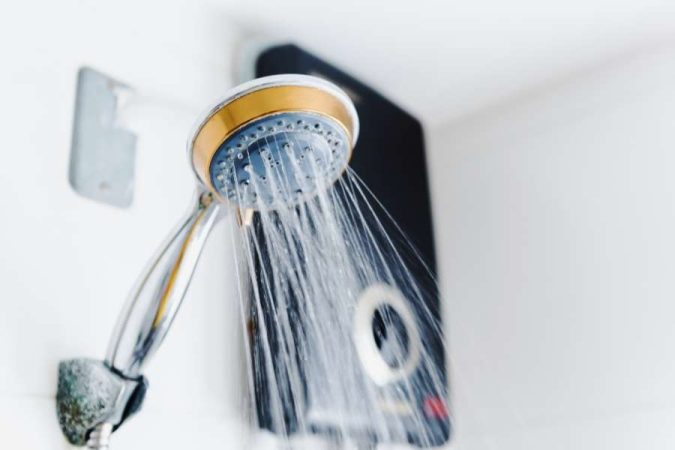 Une pomme de douche à main en fonctionnement avec un chauffe-eau électrique instantané en arrière-plan, fournissant de l'eau chaude sanitaire.